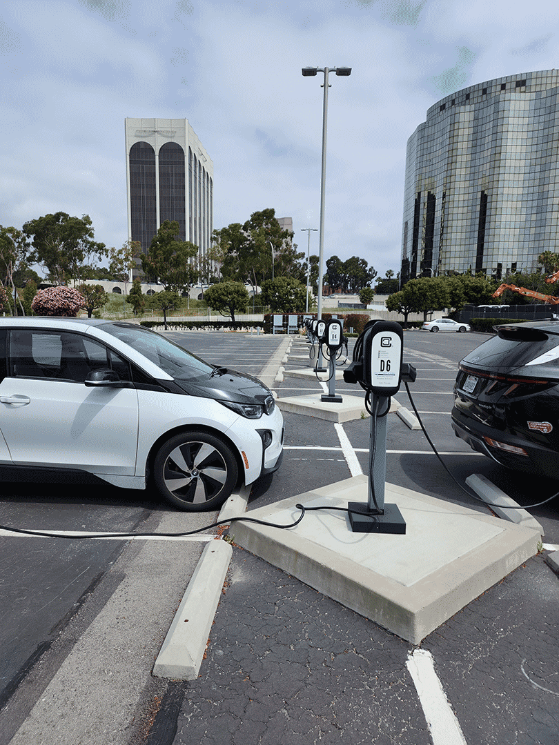 several electric vehichles charging at clipper creek or enphase EVSE 40A charge stations. Charge stations are managed by Liberty Plugins Charge Station Management System (CSMS) based on Hydra. Tall buildings are visible beyond parking lot.