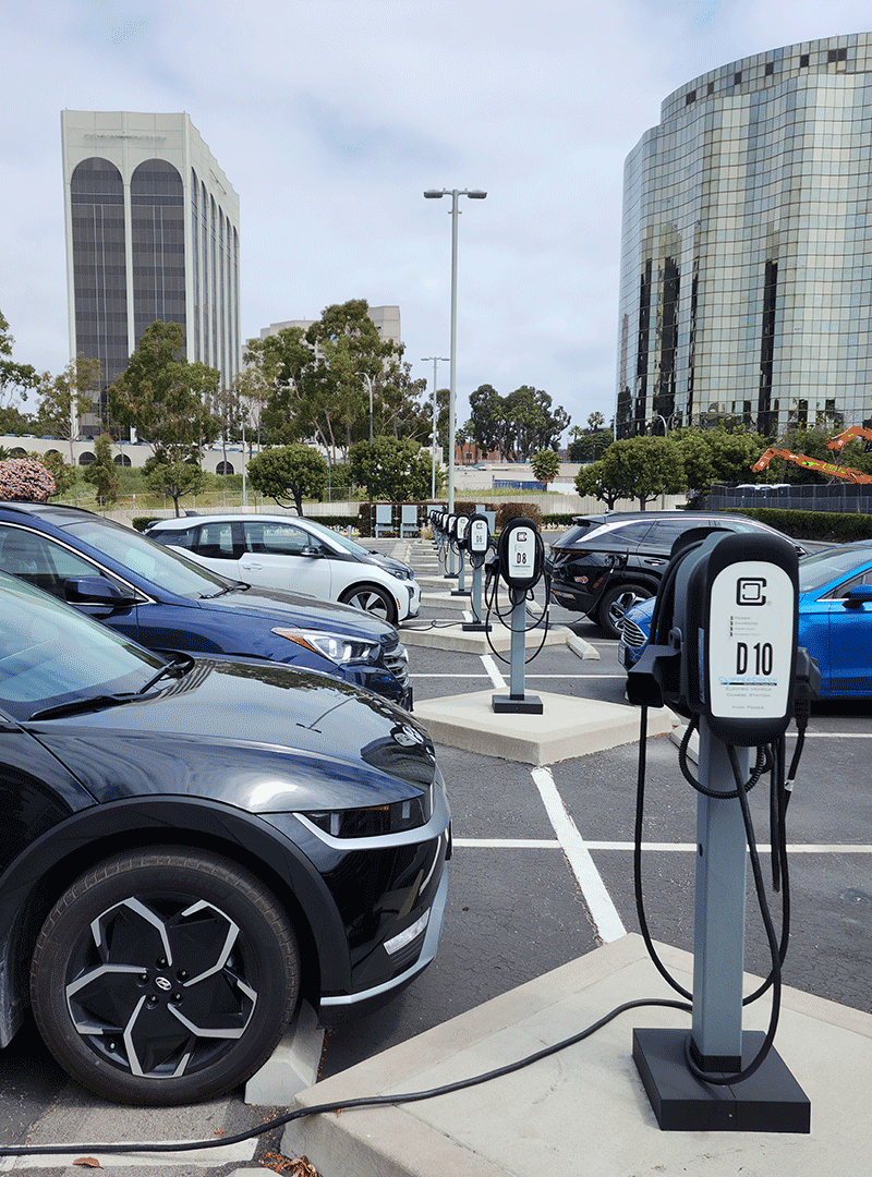 several electric vehichles charging at clipper creek or enphase EVSE 40A charge stations. Charge stations are managed by Liberty Plugins Charge Station Management System (CSMS) based on Hydra. Tall buildings are visible beyond parking lot.